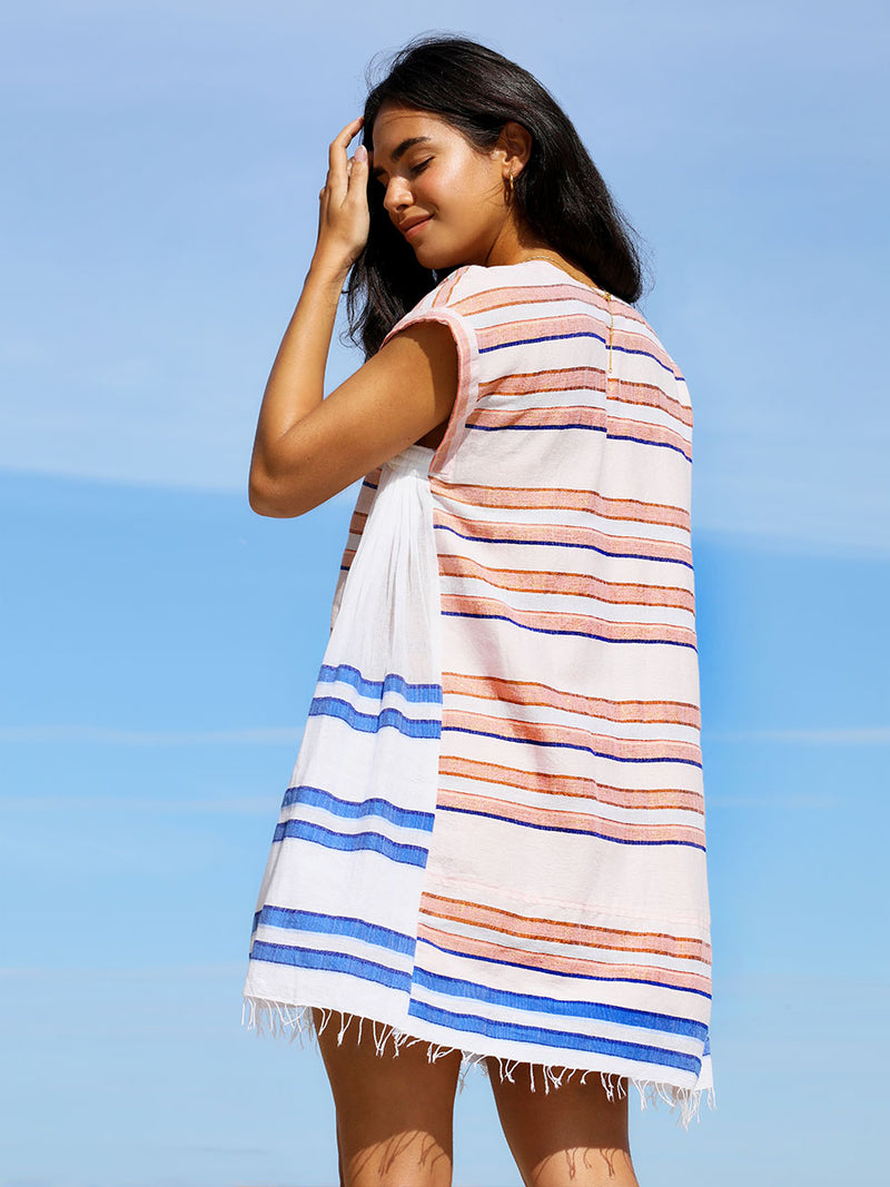 The back view of a woman standing wearing the Eskedar Caftan Dress in Seashell featuring blue, white, brown, nude and gold lurex stripes