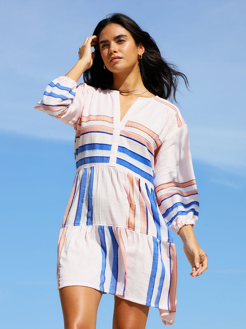 A woman standing wearing the Eskedar Popover Dress in Seashell featuring blue, white, brown, nude and gold lurex stripes