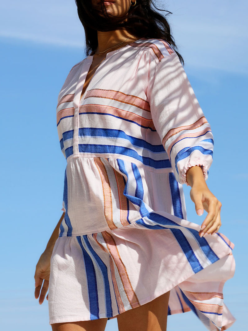 A close up of awoman standing wearing the Eskedar Popover Dress in Seashell featuring blue, white, brown, nude and gold lurex stripes