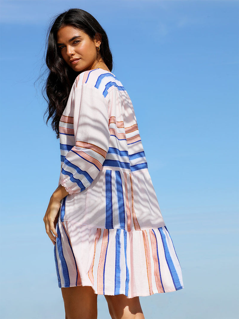 The side view of a woman standing wearing the Eskedar Popover Dress in Seashell featuring blue, white, brown, nude and gold lurex stripes