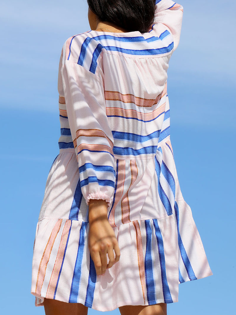 The back view of a woman standing wearing the Eskedar Popover Dress in Seashell featuring blue, white, brown, nude and gold lurex stripes