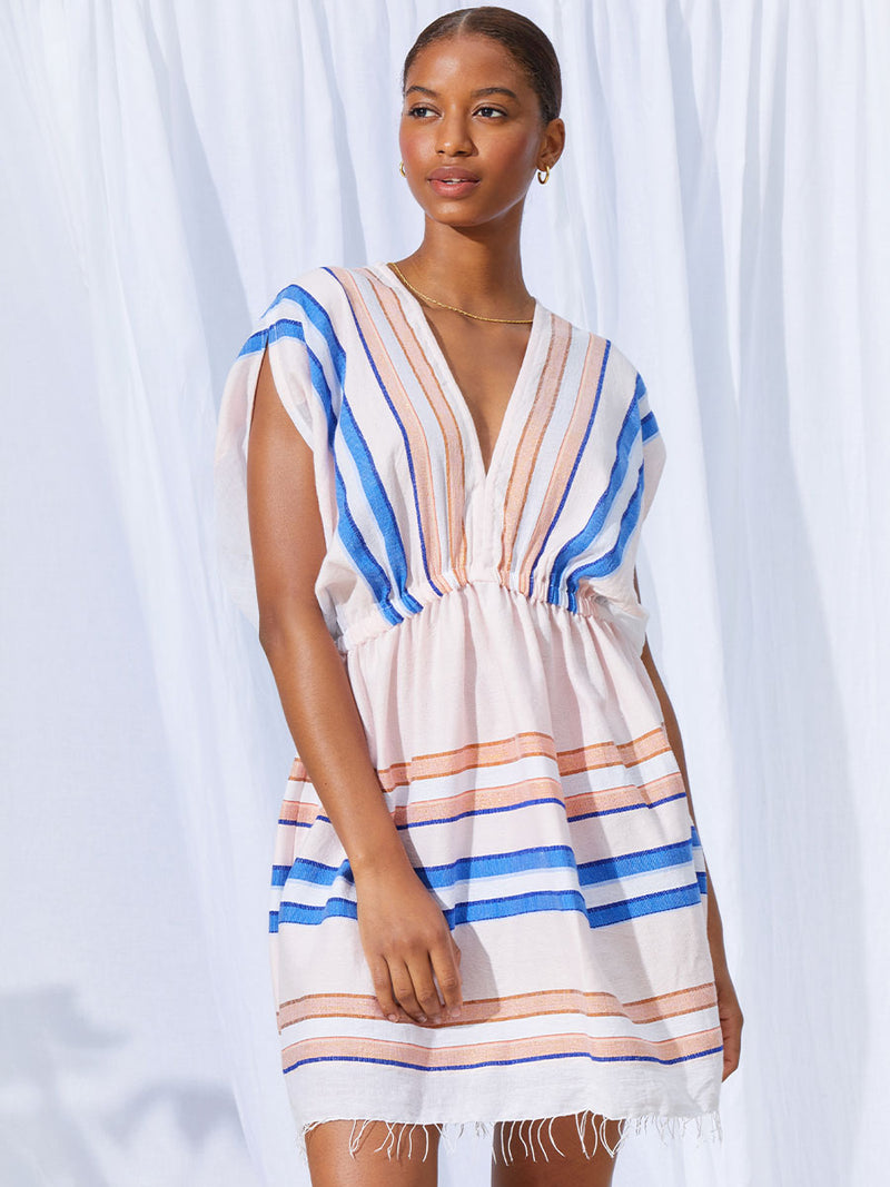 A woman standing wearing the Eskedar Short Plunge Neck Dress in Seashell featuring blue, white, brown, nude and gold lurex stripes