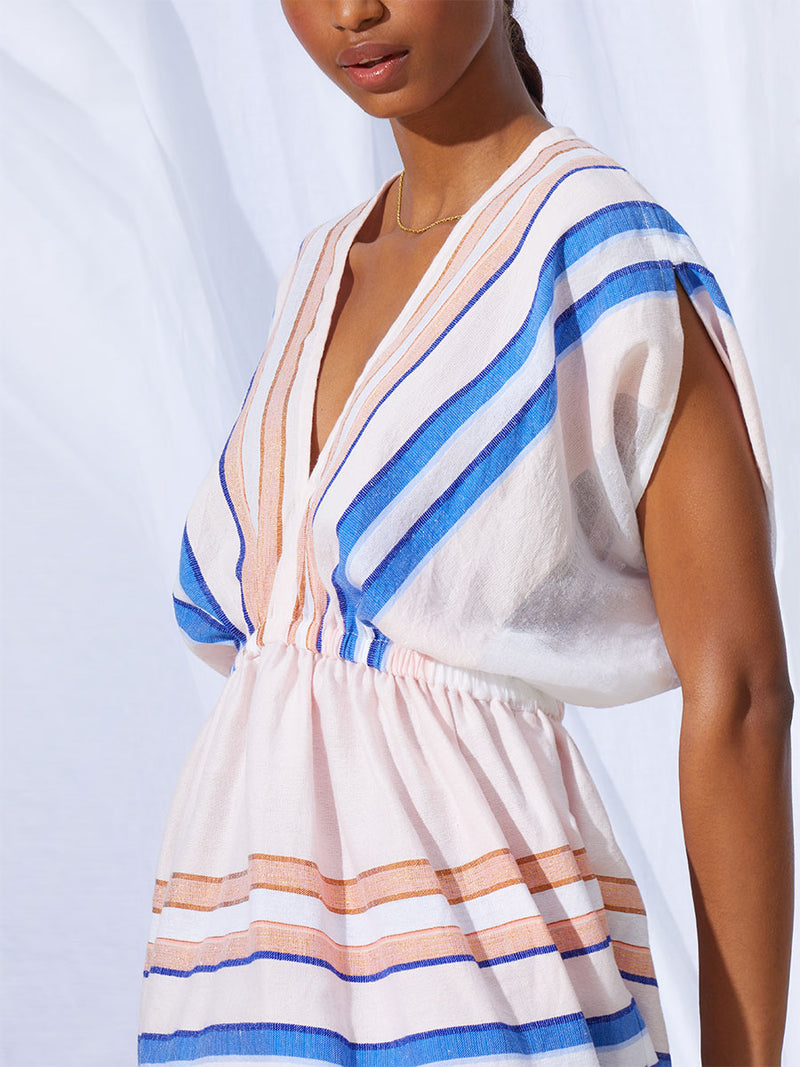 The side view of a woman standing wearing the Eskedar Short Plunge Neck Dress in Seashell featuring blue, white, brown, nude and gold lurex stripes