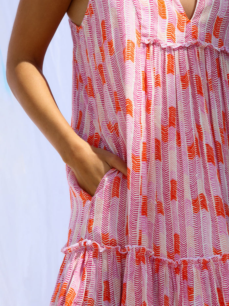The close up view of a woman standing with her hand in her pocket wearing the Gigi Bib Dress in Multi Pink featuring pink, peach and orange allover chevron print and strips of gold lurex.