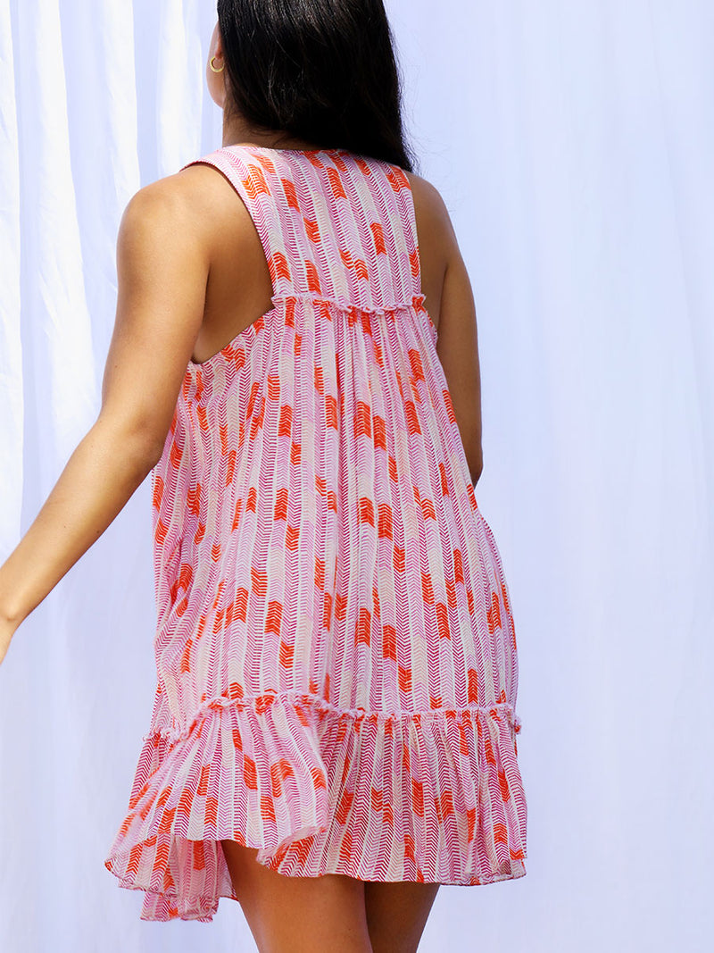 The back view of a woman walking wearing the Gigi Bib Dress in Multi Pink featuring pink, peach and orange allover chevron print and strips of gold lurex.