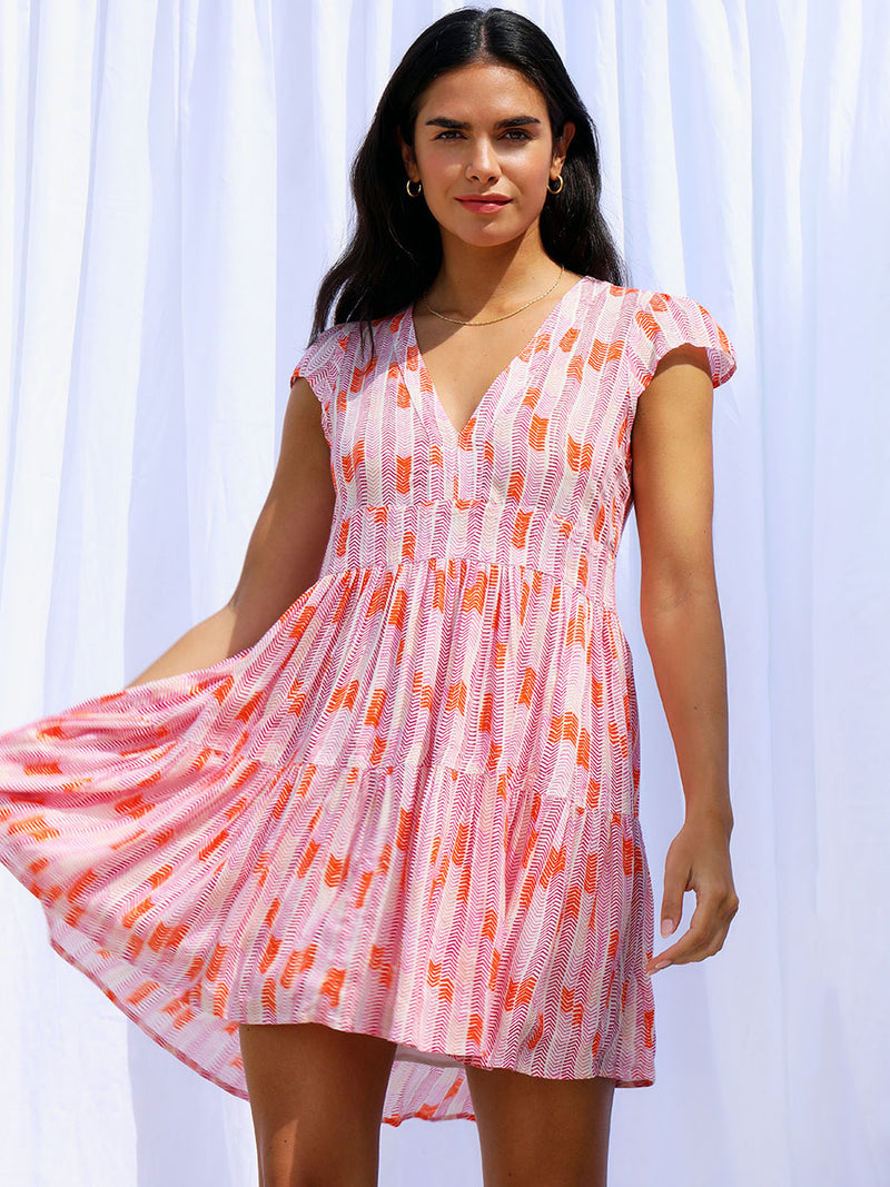 A woman standing wearing the Gigi Flutter Sleeve Dress in Multi Pink featuring pink, peach and orange allover chevron print and strips of gold lurex. 
