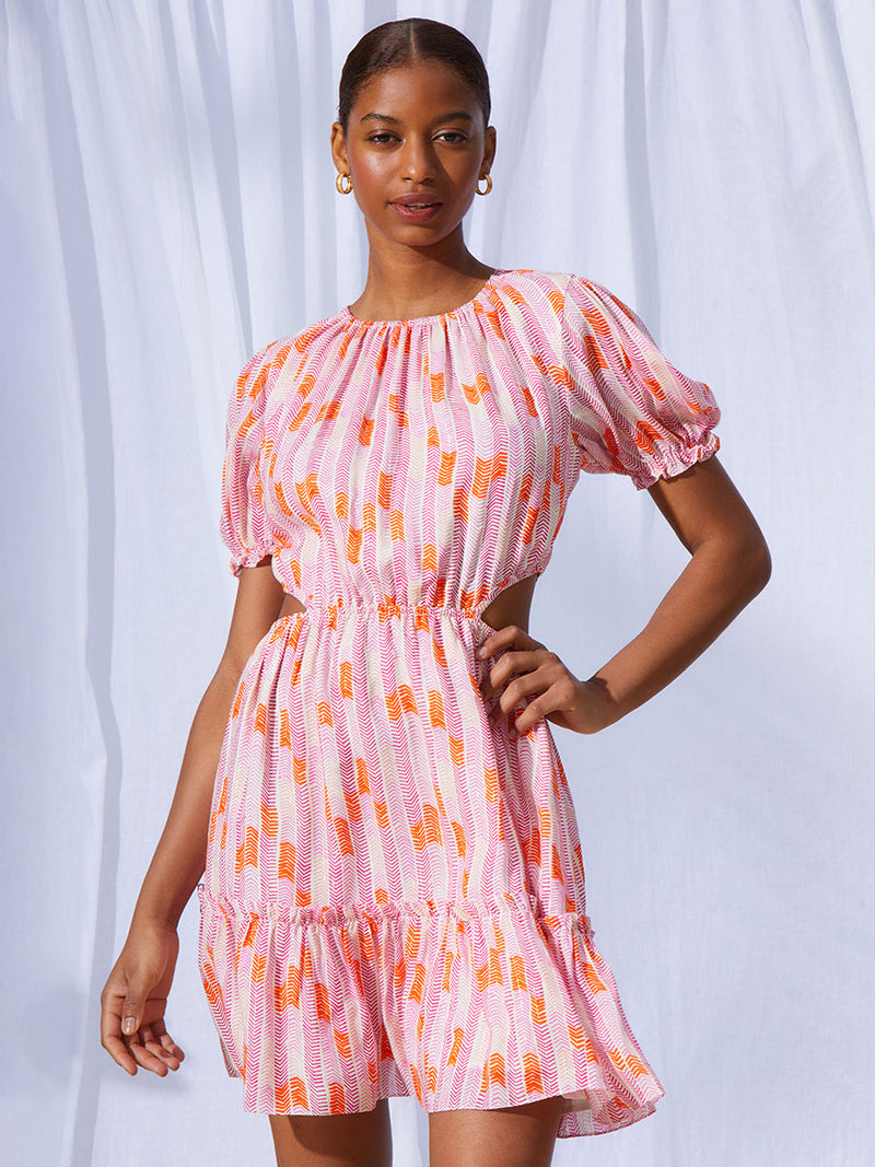 A view of a woman wearing the Gigi Short Open Back Dress in Multi Pink featuring pink, peach and orange allover chevron print and strips of gold lurex.