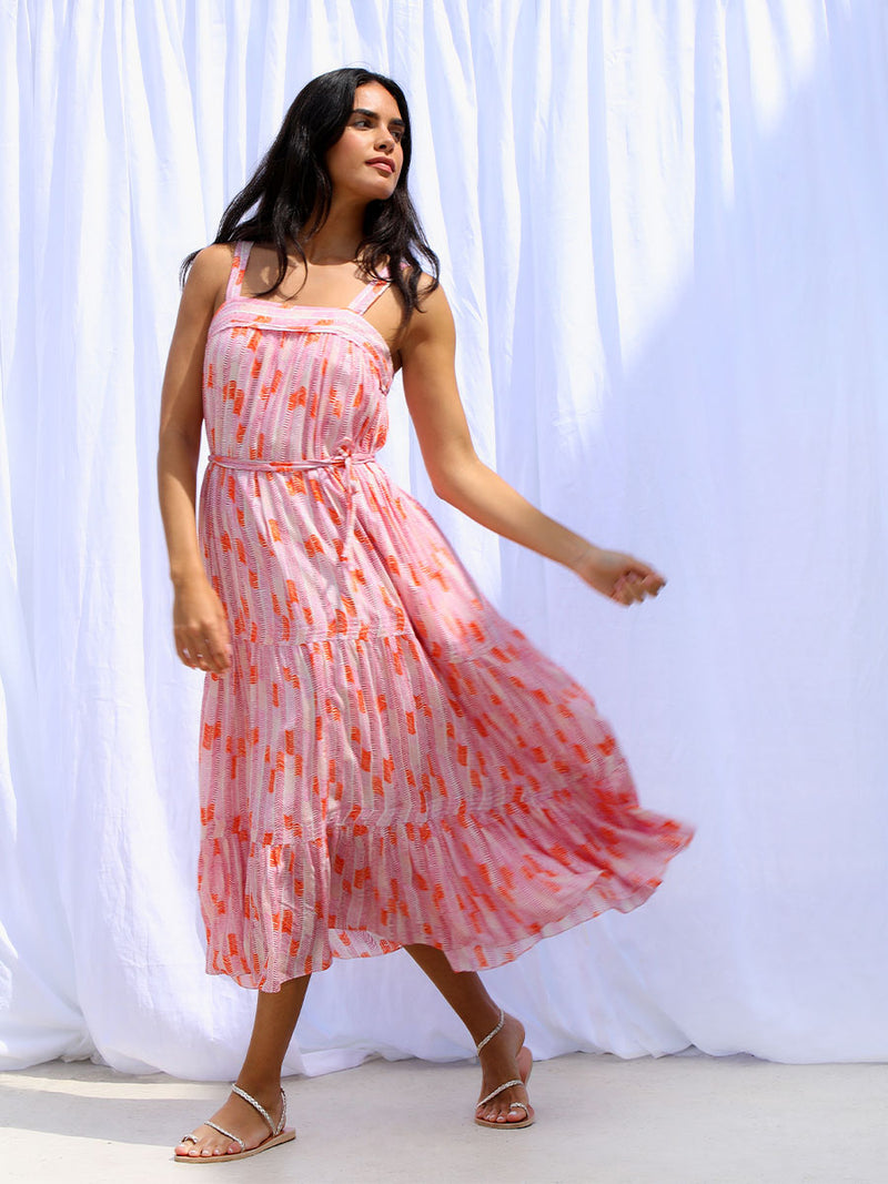 A woman standing wearing the Gigi Sweepy Sundress in Multi Pink featuring pink, peach and orange allover chevron print and strips of gold lurex. 