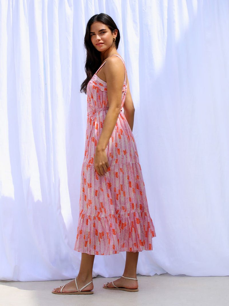 A side view of a woman standing wearing the Gigi Sweepy Sundress in Multi Pink featuring pink, peach and orange allover chevron print and strips of gold lurex.