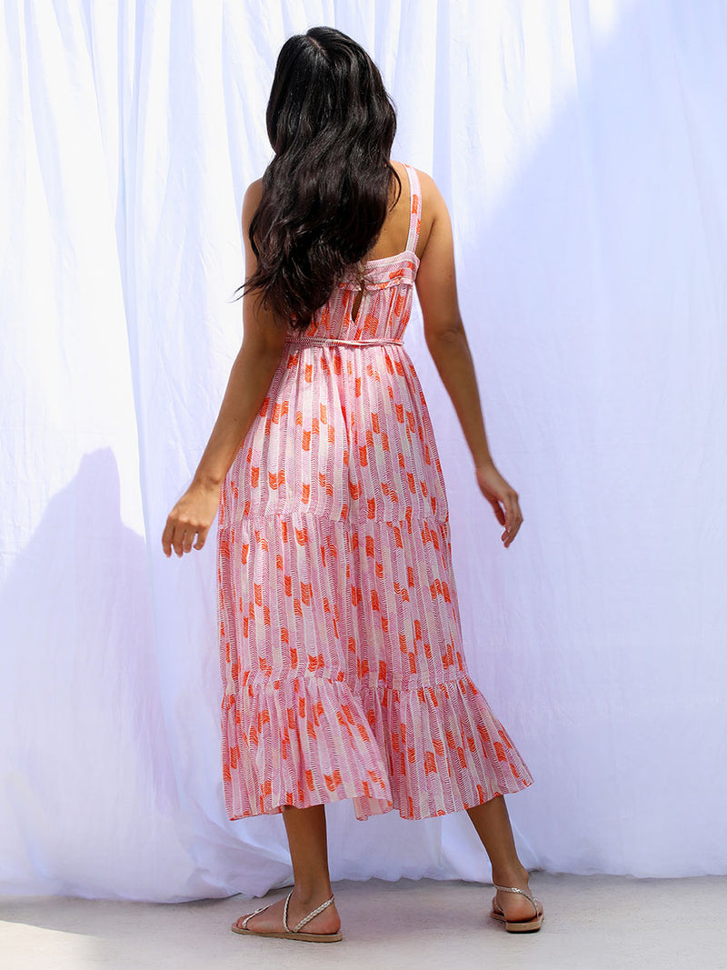 A back view of a woman standing wearing the Gigi Sweepy Sundress in Multi Pink featuring pink, peach and orange allover chevron print and strips of gold lurex.