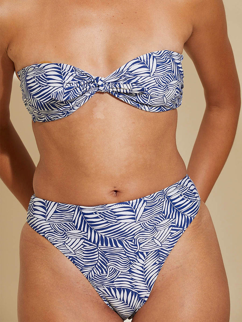 Woman standing with her arms behind her back wearing the Palm Leaf Bandeau Top and matching High Leg Bikini Bottom featuring palm tree patterns on a rich blue background.