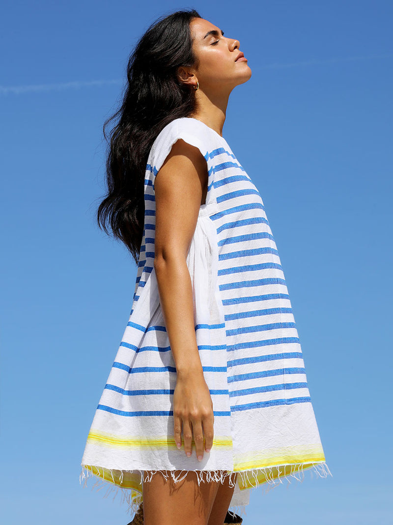 A side view of a woman standing wearing the Hirut Caftan Dress in Blue featuring blue stripes on white foreground and yellow degrade stripes at the bottom hem.