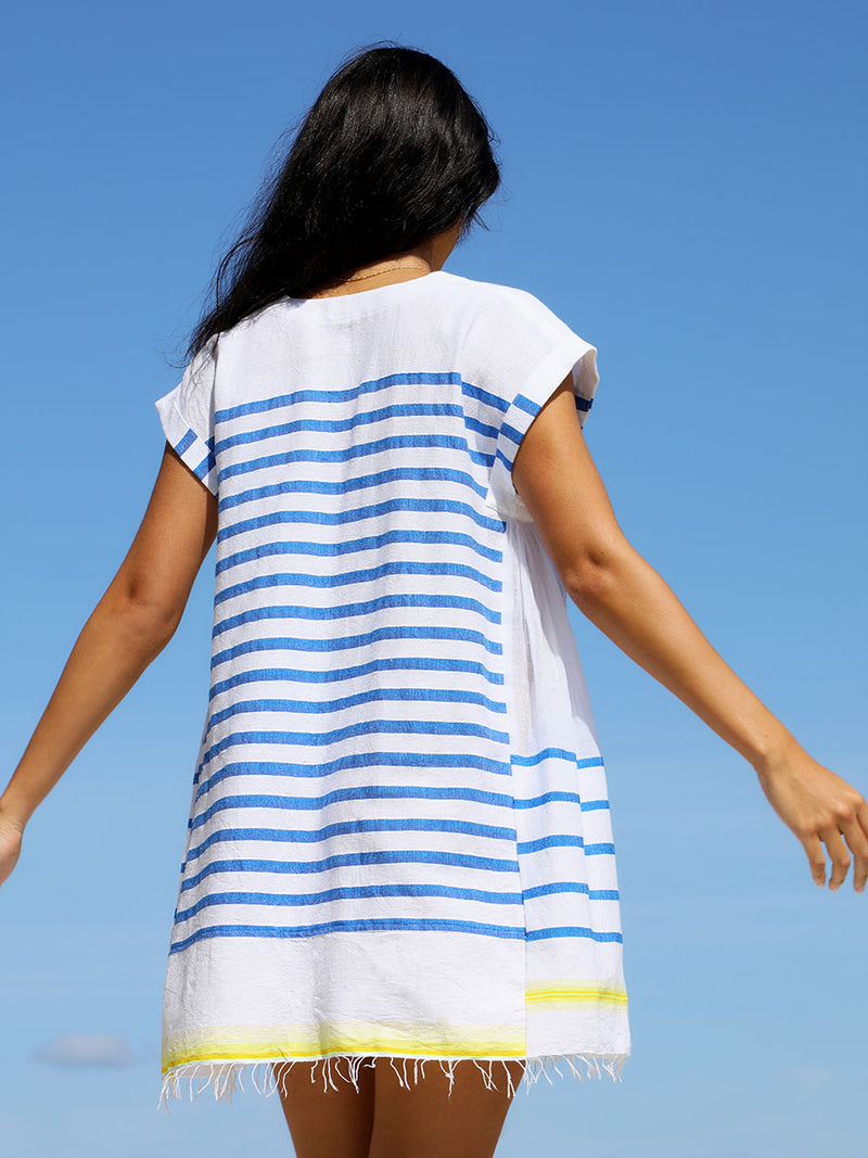 A back view of a woman standing wearing the Hirut Caftan Dress in Blue featuring blue stripes on white foreground and yellow degrade stripes at the bottom hem.