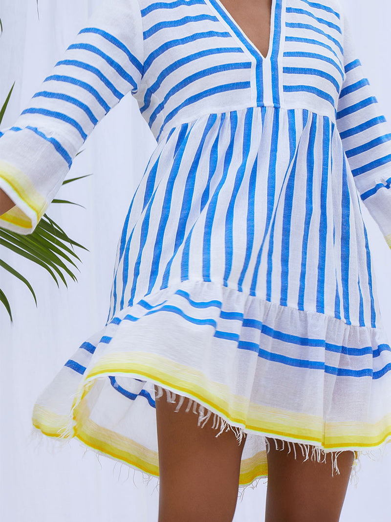 A close up view of a woman standing wearing the Hirut Flutter Dress in Blue featuring blue stripes on white foreground and yellow degrade stripes at the edges.