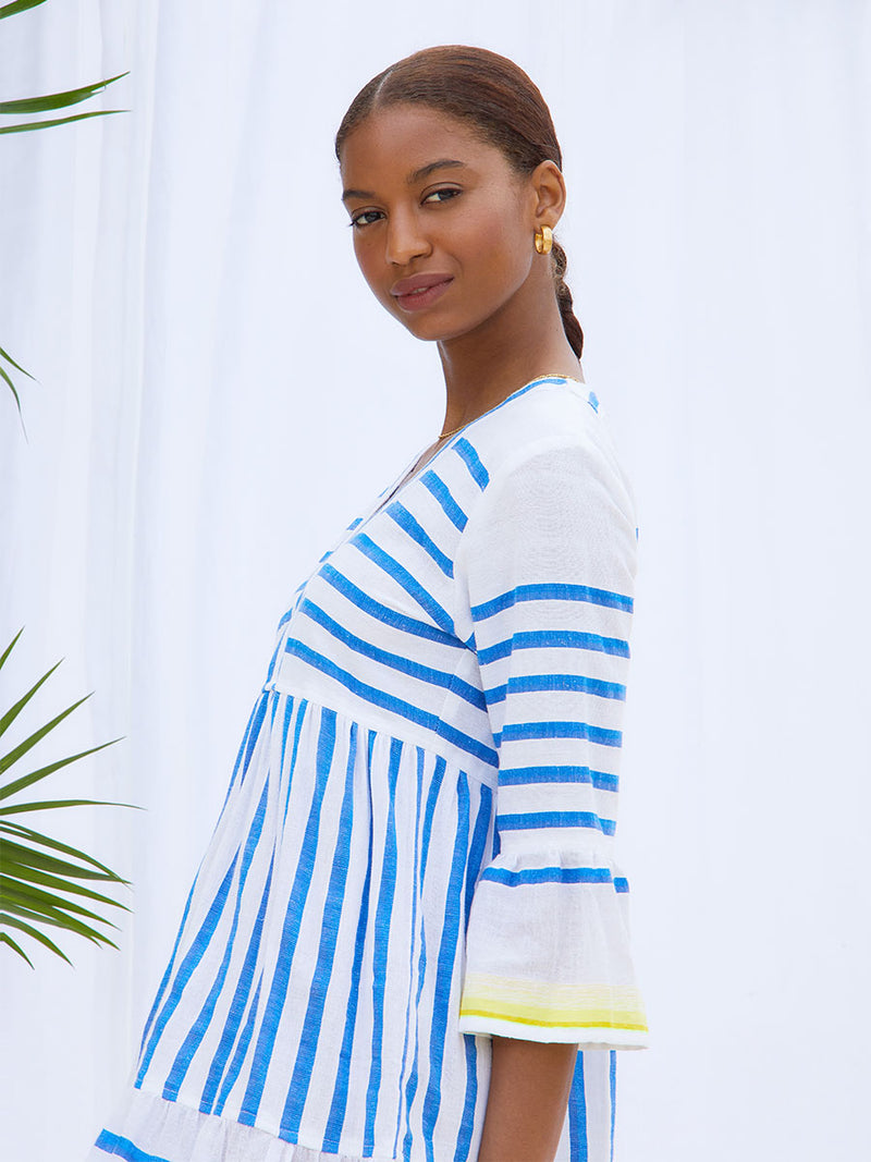 A side view of a woman standing wearing the Hirut Flutter Dress in Blue featuring blue stripes on white foreground and yellow degrade stripes at the edges.