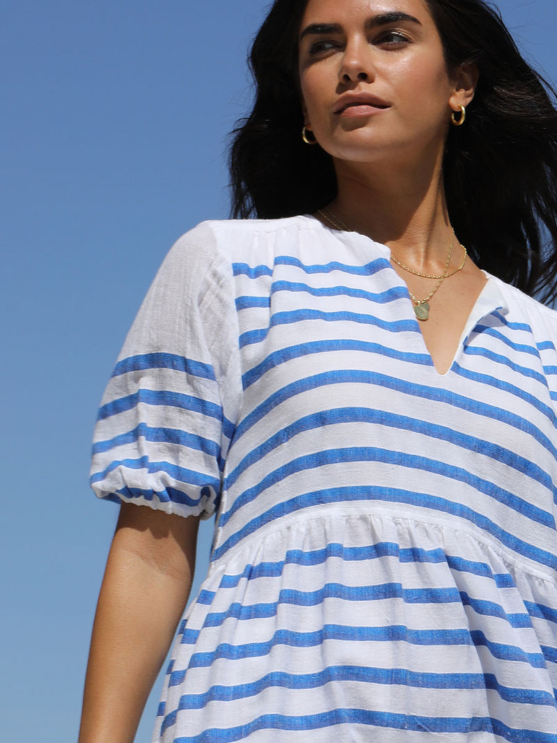 Woman standing looking over her shoulder wearing the Hirut Popover Dress in Blue featuring blue stripes on white foreground and yellow degrade stripes across the top back and bottom hem.
