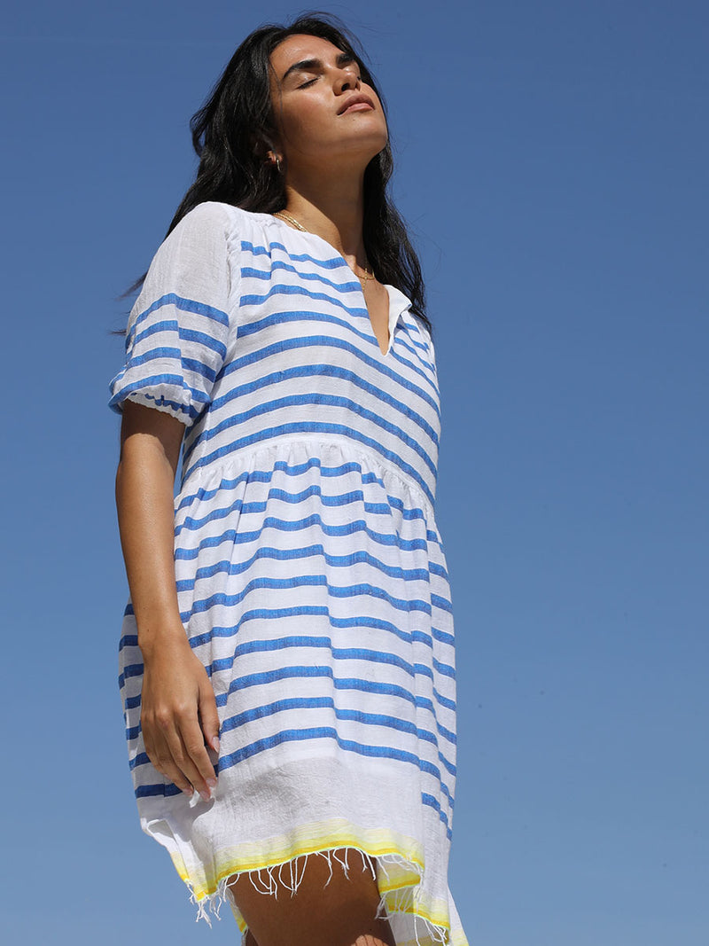 Woman standing with the eyes closed wearing the Hirut Popover Dress in Blue featuring blue stripes on white foreground and yellow degrade stripes across the top back and bottom hem.