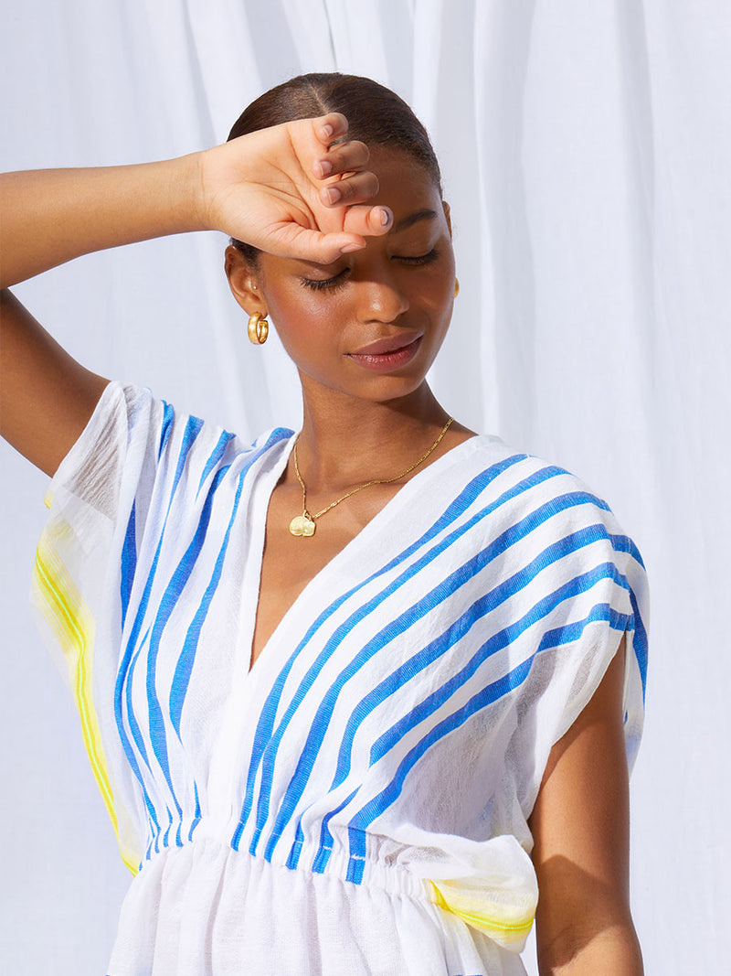 A close up of woman standing with her hands on her face wearing the Hirut Short Plunge Neck Dress in Blue featuring blue stripes on white foreground and yellow degrade stripes at the edges.