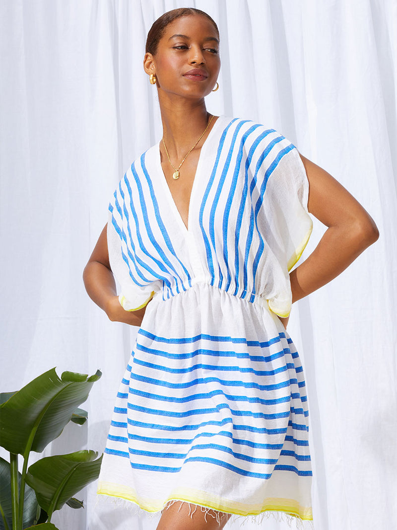 A woman standing with her hands on her hips wearing the Hirut Short Plunge Neck Dress in Blue featuring blue stripes on white foreground and  yellow degrade stripes at the edges.