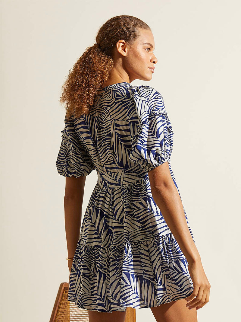 Back view of a woman standing wearing the Palm Leaf Flutter Sleve Mini Dress featuring palm tree patterns on a rich blue background.