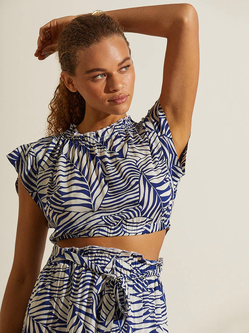 Woman standing with her hand over her head wearing the Palm Leaf Maxi Skirt and matching Ruched Crop Topfeaturing palm tree patterns on a rich blue background.
