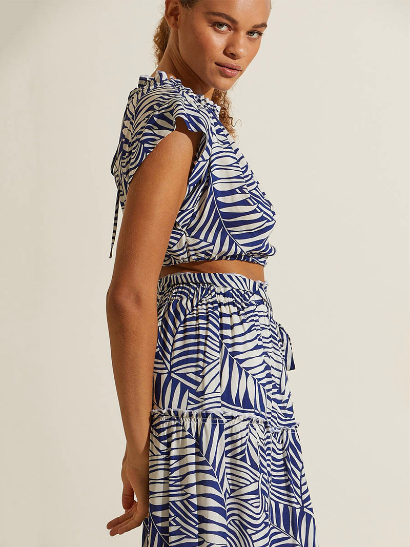 Side view of a woman standing with her hand in her back wearing the Palm Leaf Maxi Skirt and matching Ruched Crop Top featuring palm tree patterns on a rich blue background.