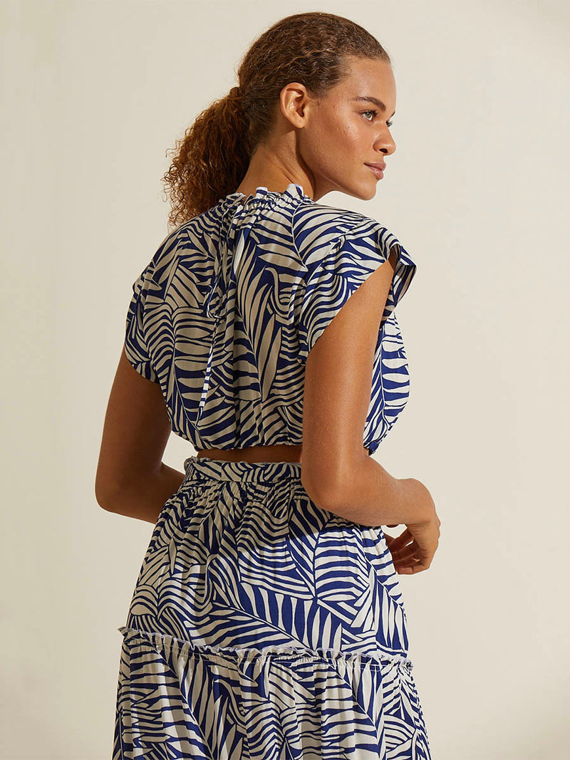 Back view a woman standing wearing the Palm Leaf Maxi Skirt and matching Ruched Crop Topfeaturing palm tree patterns on a rich blue background.