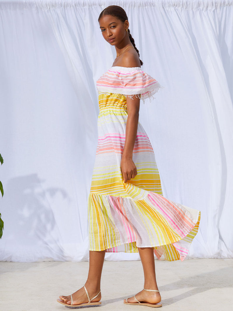 A side view of a woman standing wearing the Jamila Beach Dress featuring shades of yellow and pink stripes on white foreground.