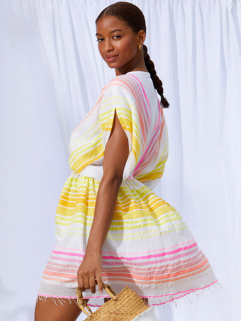 A side view of a woman standing wearing the Jamila Short Plunge Dress featuring shades of yellow and pink stripes on white foreground.