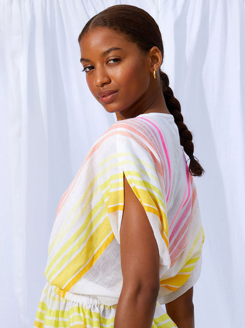 A close up on the side of woman standing wearing the Jamila Short Plunge Dress featuring shades of yellow and pink stripes on white foreground.