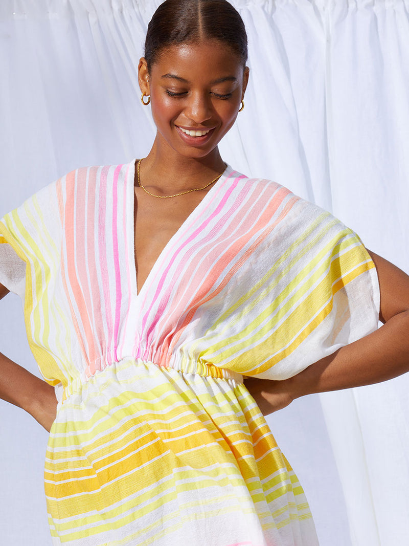 A view woman standing with her hands on her hips wearing the Jamila Short Plunge Dress featuring shades of yellow and pink stripes on white foreground.