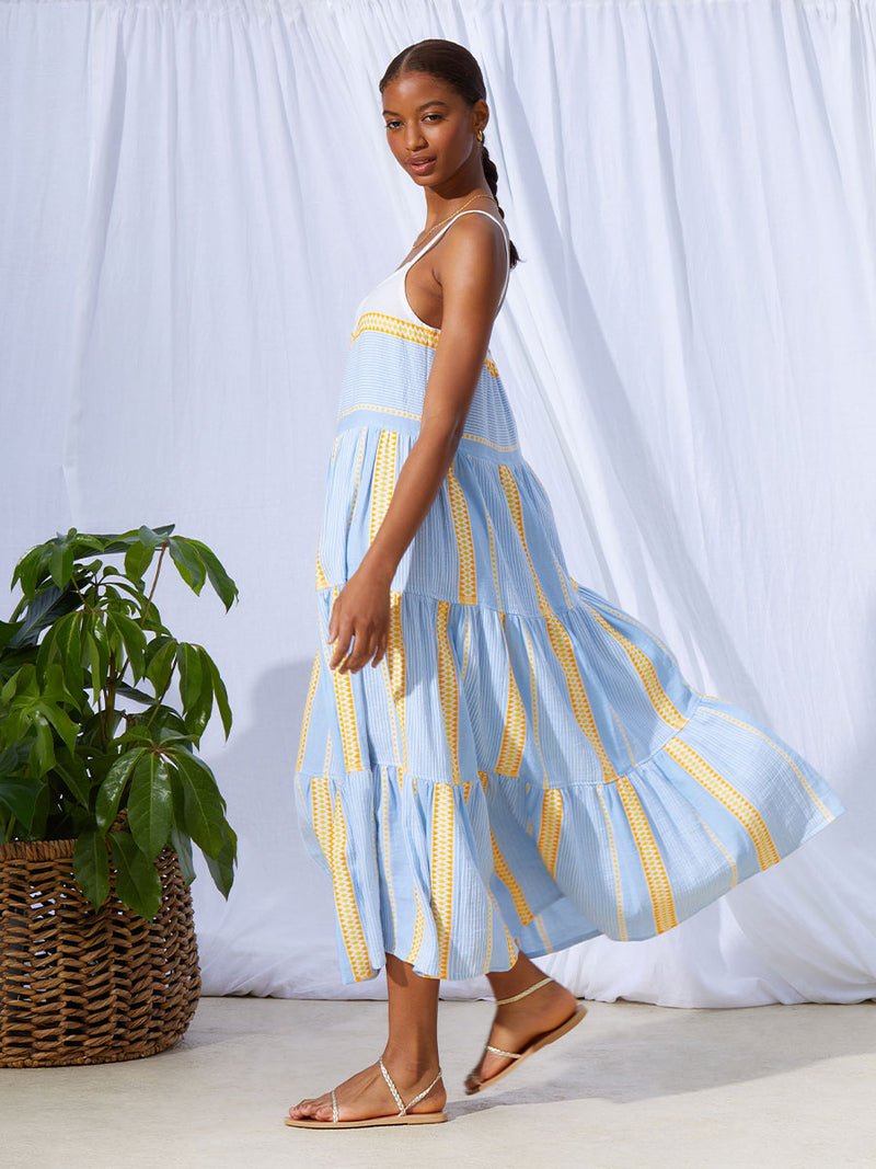 The side view of a woman standing wearing the Jemari Cascade Dress in sky blue featuring yellow and orange diamond patterns