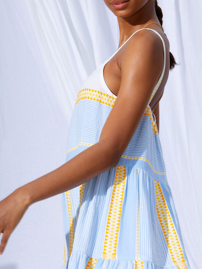 A close up view of a woman standing wearing the Jemari Cascade Dress in sky blue featuring yellow and orange diamond patterns