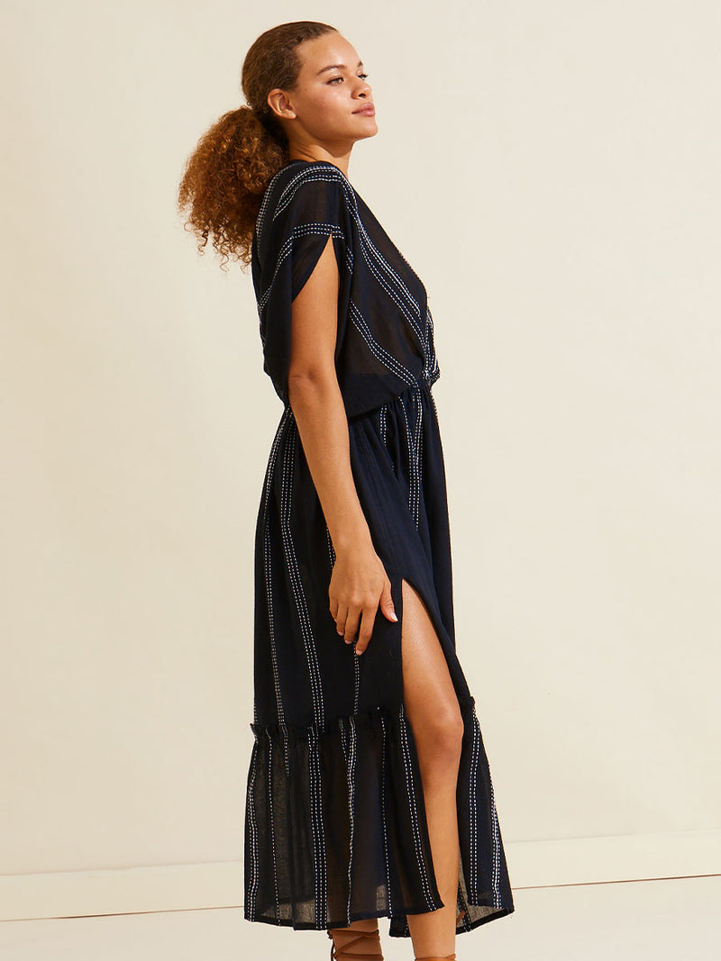 Side view of a woman standing wearing the Leliti Plunge Neck Dress in Black with white stitching allover.