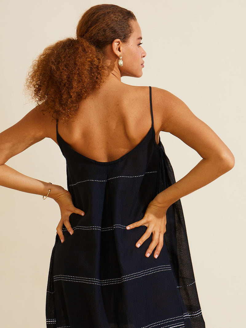 Back view of a woman standing with her hands on her hips wearing the Leliti Slip Dress in Black with white stitching allover.