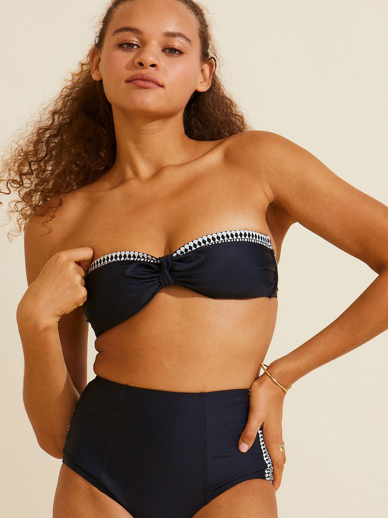 Woman standing with one hand on her hips and the other by her chest wearing the Lena Bandeau bikini top and matching High Waist Bottom in black featuring a black and white tibeb trim.