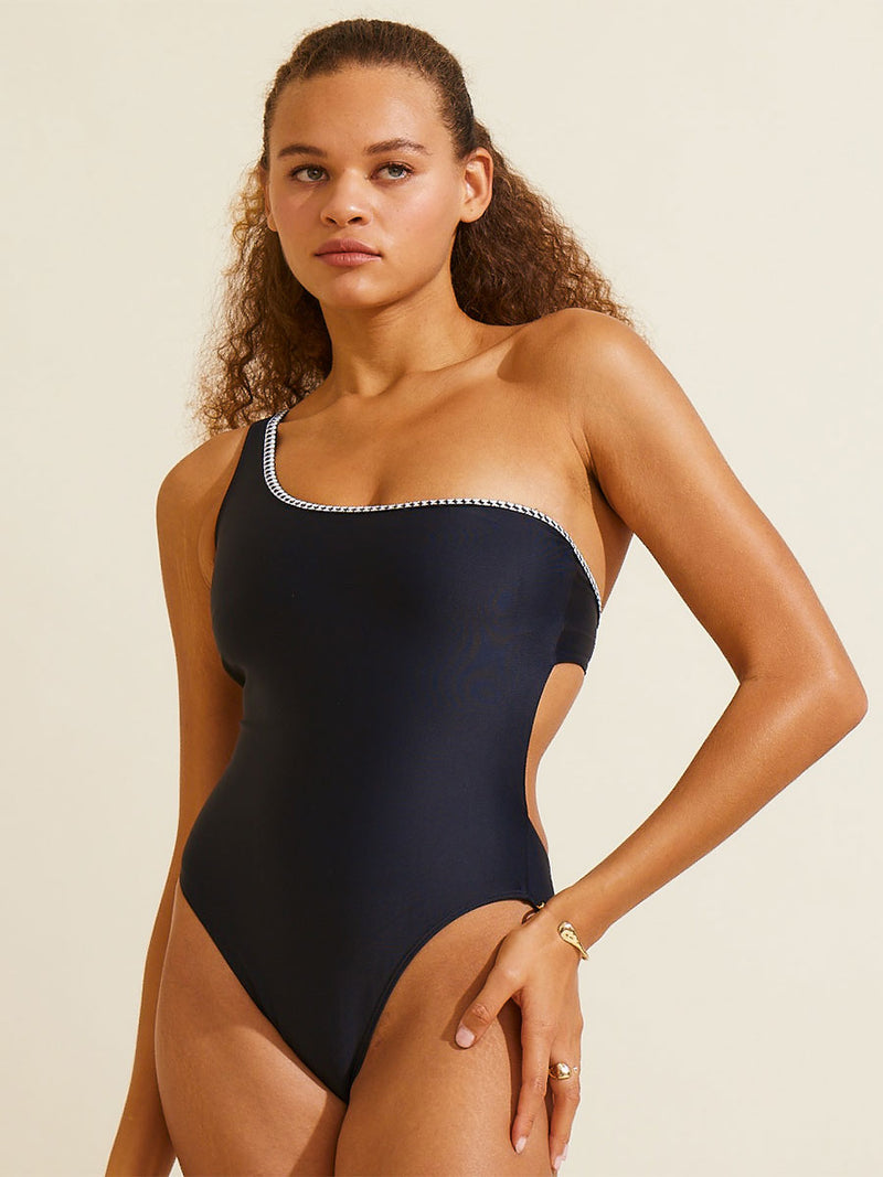 A woman standing with one hand on her hips wearing the Lena One Shoulder One Piece in Black featuring a black and white tibeb trim.