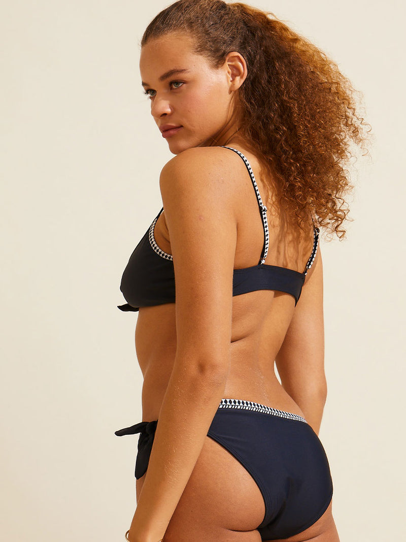 Side view of a woman standing wearing the lena tie front bikini top and matching sofia side tie bikini bottom in black with graphic white diamond trim