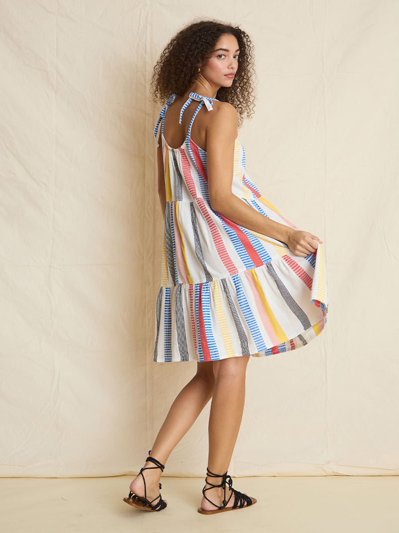 The back view of a woman standing wearing the Meseret Midi Cascade Dress featuring a variety of pink, yellow, black, red, and blue stripe patterns. 