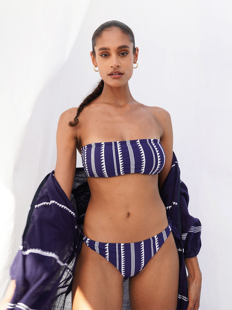 Woman wearing a navy Nunu bandeau top with triangles and stripes with matching side tie bikini bottom and long robe