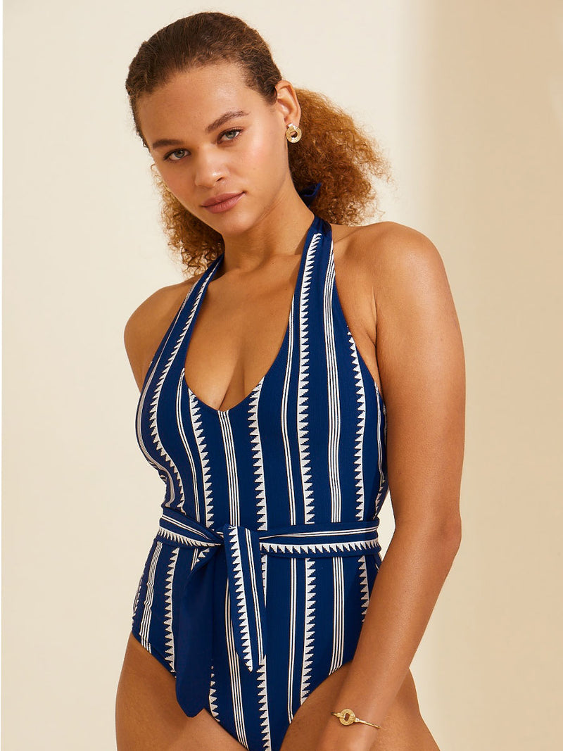 Woman standing wearing a navy Nunu deep v belted one piece swimsuit with white triangles and stripes