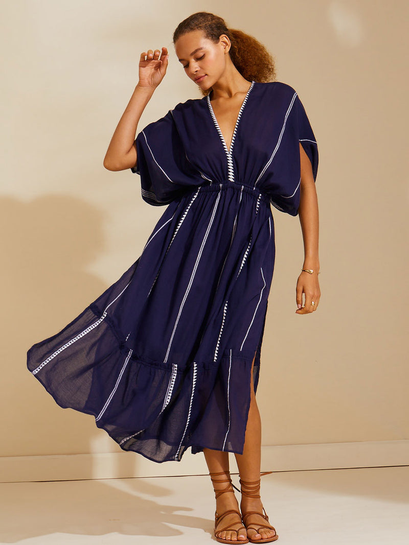 Woman standing with her hand by her head wearing  the Nunu Plunge Neck dress in navy blue featuring a navy with white vertical stripes and graphic lines.