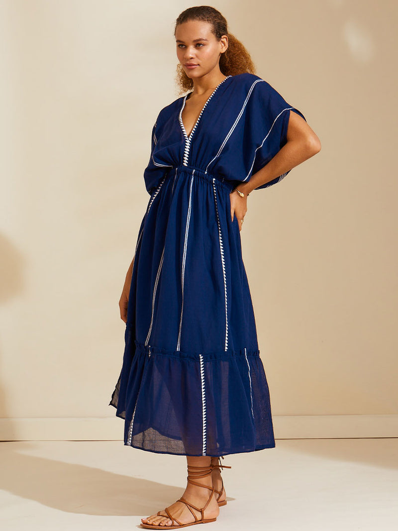 Side view of a woman standing with her hand on her hips wearing  the Nunu Plunge Neck dress in navy blue featuring a navy with white vertical stripes and graphic lines.