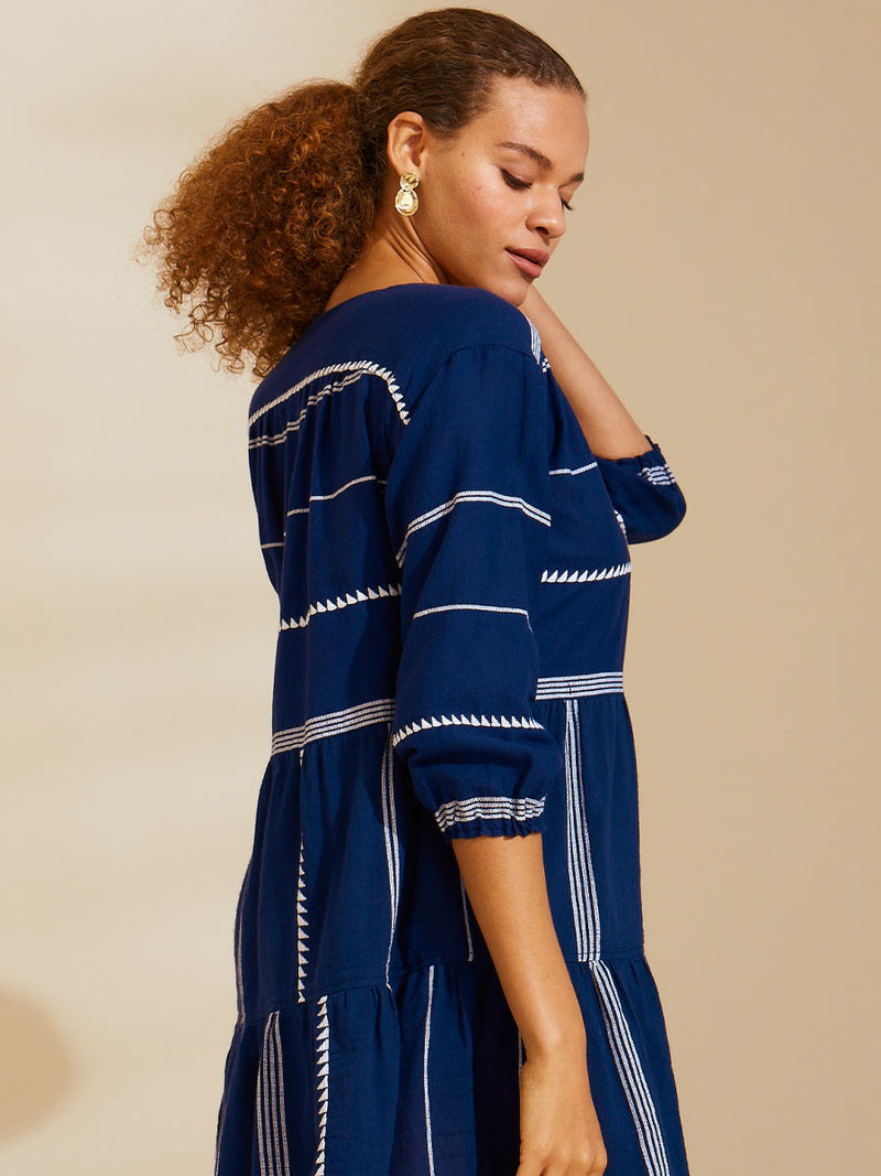 Side view of a woman standing with her hand on her head wearing navy Nunu short popover dress with white triangles and stripes