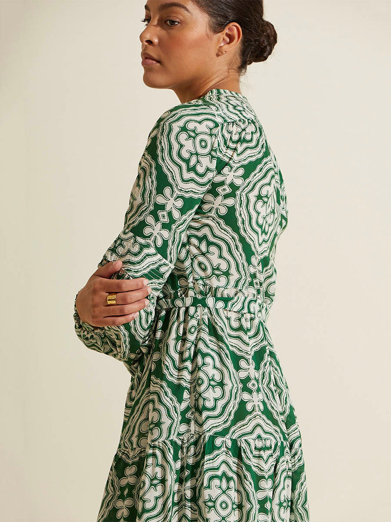 Side view of a woman standing with her arms crossed wearing the Medallion Peasant Dress featuring architectural white patterns on a deep green background.