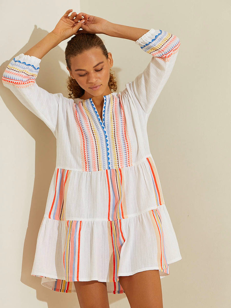 Woman standing with her hands over her head wearing the Bekah Popover Dress featuring 10 tutti frutti colors embroidered on a white background.  