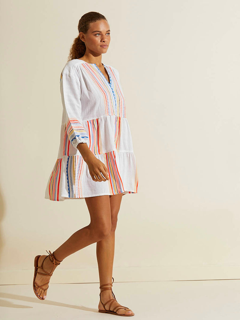 Side view of a woman wearing the Bekah Popover Dress featuring 10 tutti frutti colors embroidered on a white background.  