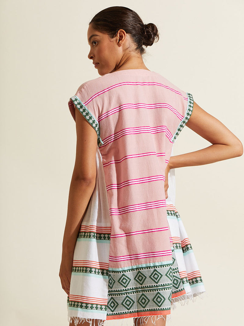 Woman standing with her hand on her hip wearing the Rosa Caftan Dress featuring architectural details and geometric border patterns woven on a soft pink background.
