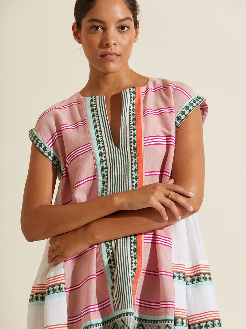 Woman standing with her arms crossed wearing the Rosa Caftan Dress featuring architectural details and geometric border patterns woven on a soft pink background.