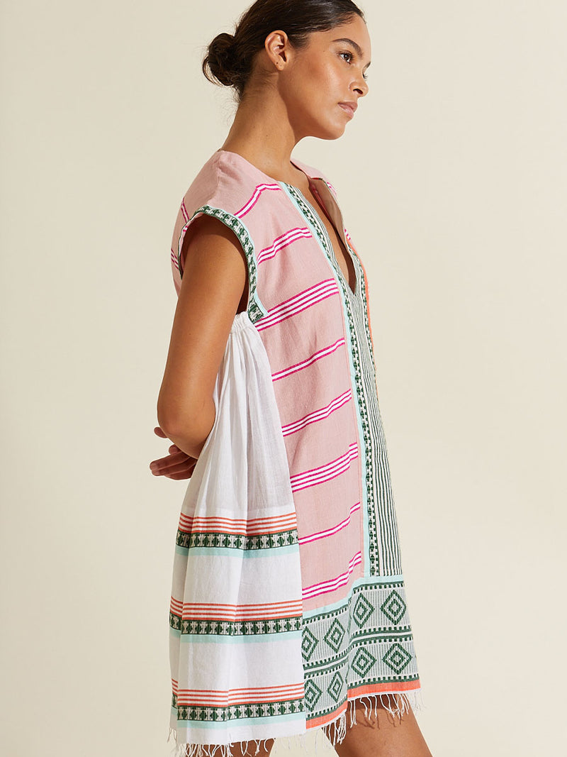 Side view of a woman standing wearing the Rosa Caftan Dress featuring architectural details and geometric border patterns woven on a soft pink background.